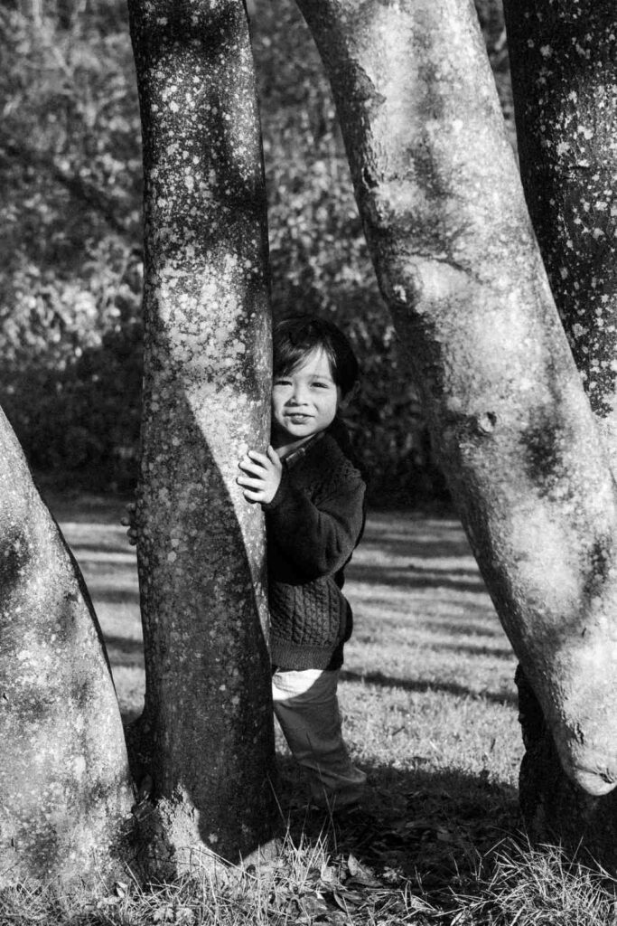 Candid black and white portrait of a child peeking from behind trees in a Vancouver park, captured during a playful family session.
